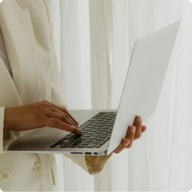 Person in white jacket holding a white laptop in front of white curtains.