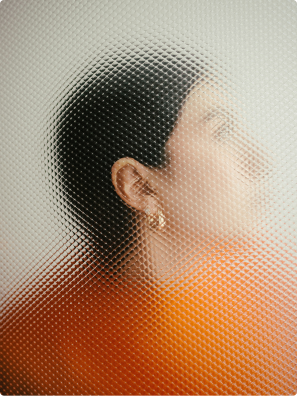 Woman in orange sweater holding her ear over frosted glass.